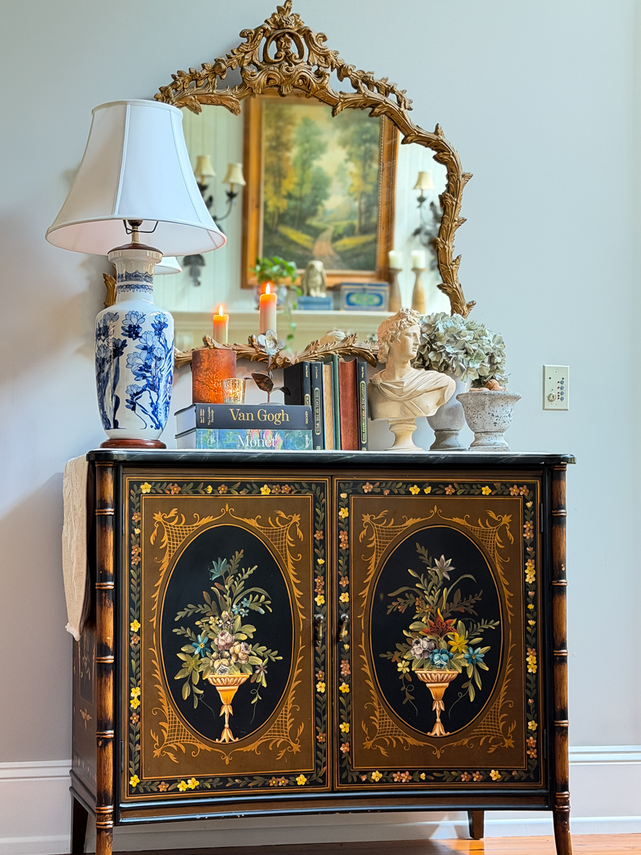 Traditional painted cabinet styled with an ornate gold mirror, blue and white lamp, vintage books, bust, and hydrangeas.