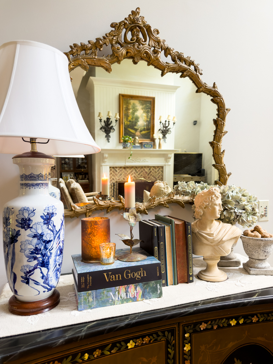 Blue and white lamp, vintage art books, candles, classical bust, and hydrangeas styled beneath an ornate gold mirror.