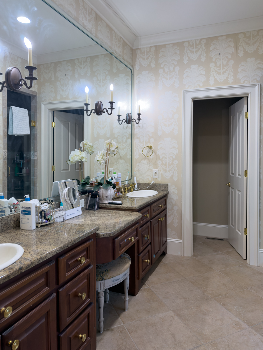 Primary bathroom with neutral patterned wallpaper, large mirror, sconces, and double vanity