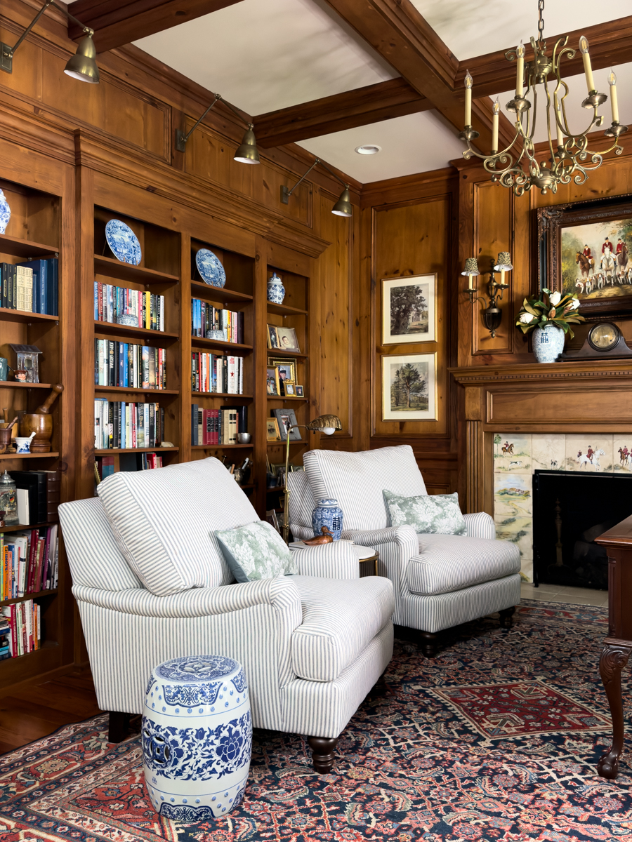 Traditional wood-paneled den with two striped chairs, built-in bookcases, fireplace, and blue and white decor