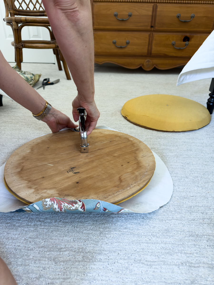 Stapling new blue floral fabric to the underside of a round wooden chair seat.