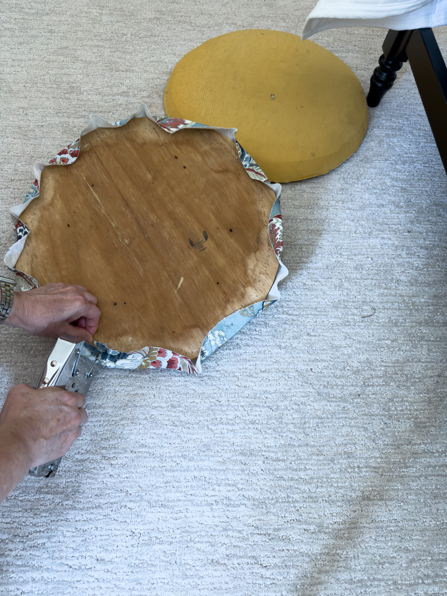 Stapling and folding blue floral fabric around the curved edge of a round wooden chair seat.