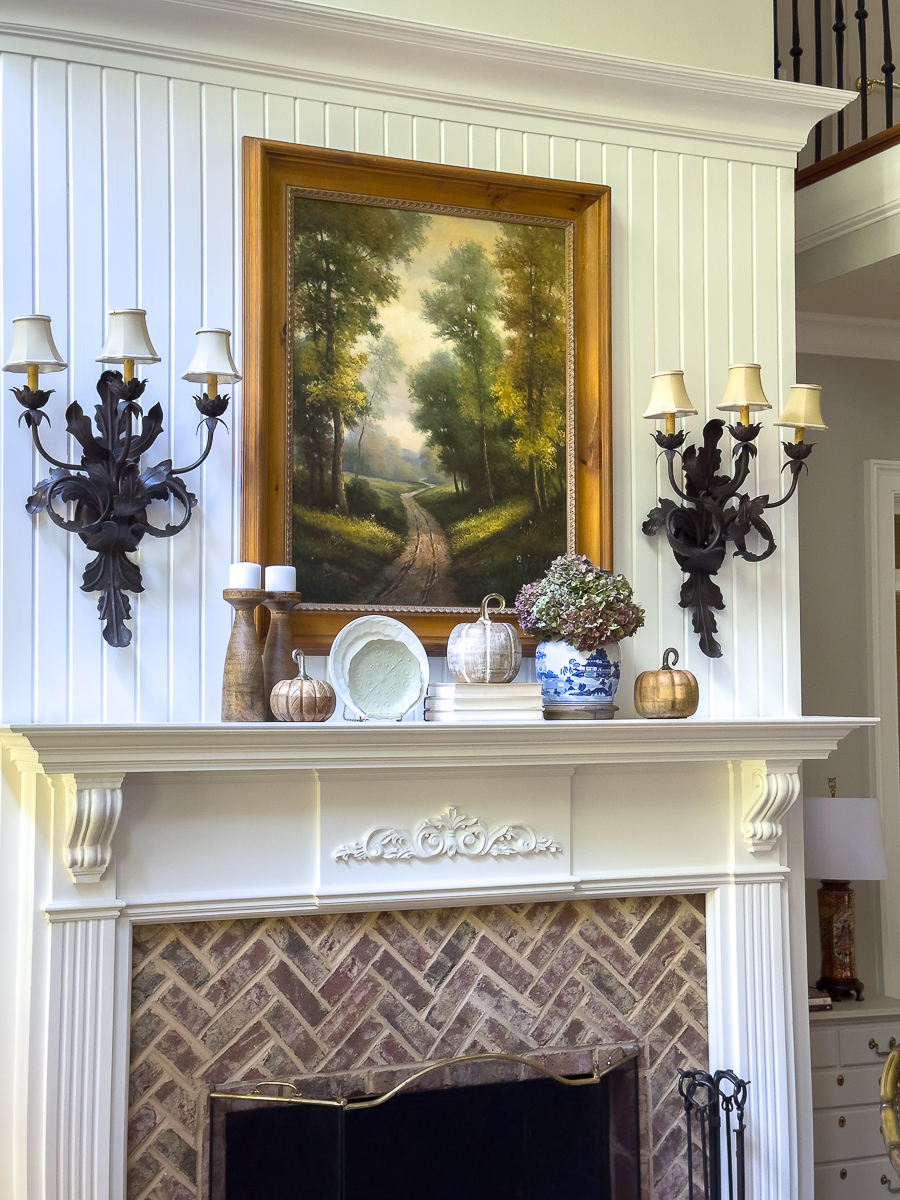 Fall mantel decorated with pumpkins, chinoiserie vase with dried hydrangeas, candles, and layered books beneath a framed landscape painting.