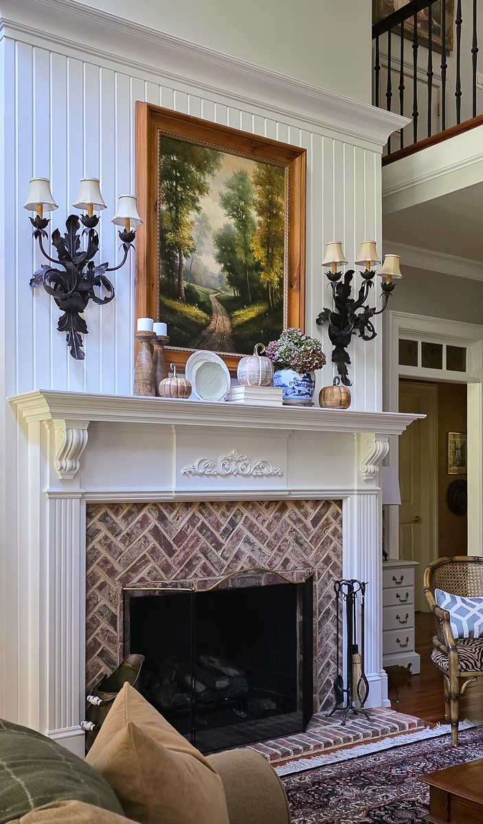 A cozy fall living room mantel with a brick fireplace, classic artwork featuring chinoiserie, pumpkins, and dried hydrangeas.