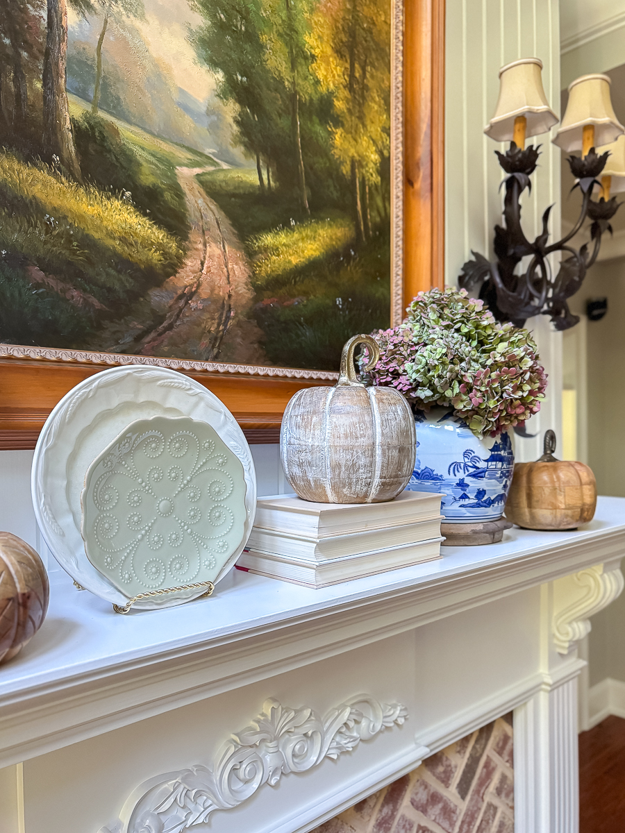 Decorative plate, stacked books, wooden pumpkins, and a chinoiserie vase with dried hydrangeas styled on a fall mantel.