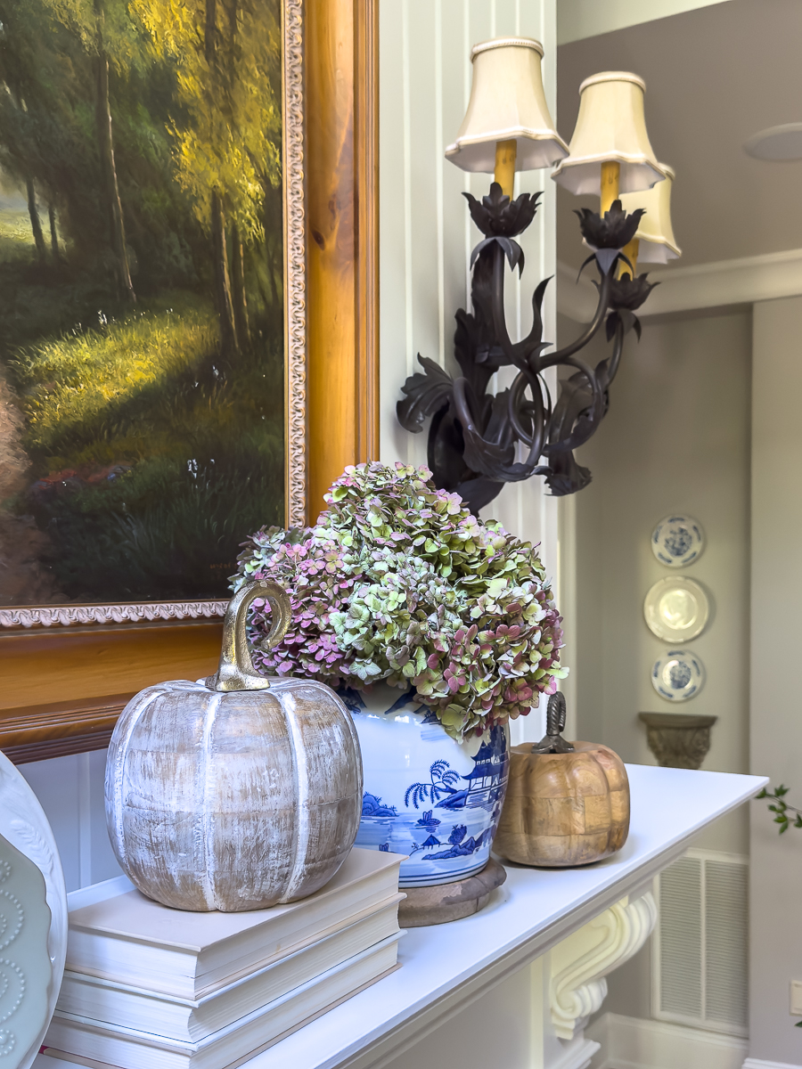 Stacked books, wooden pumpkins, and a chinoiserie vase with dried hydrangeas styled on a fall mantel.
