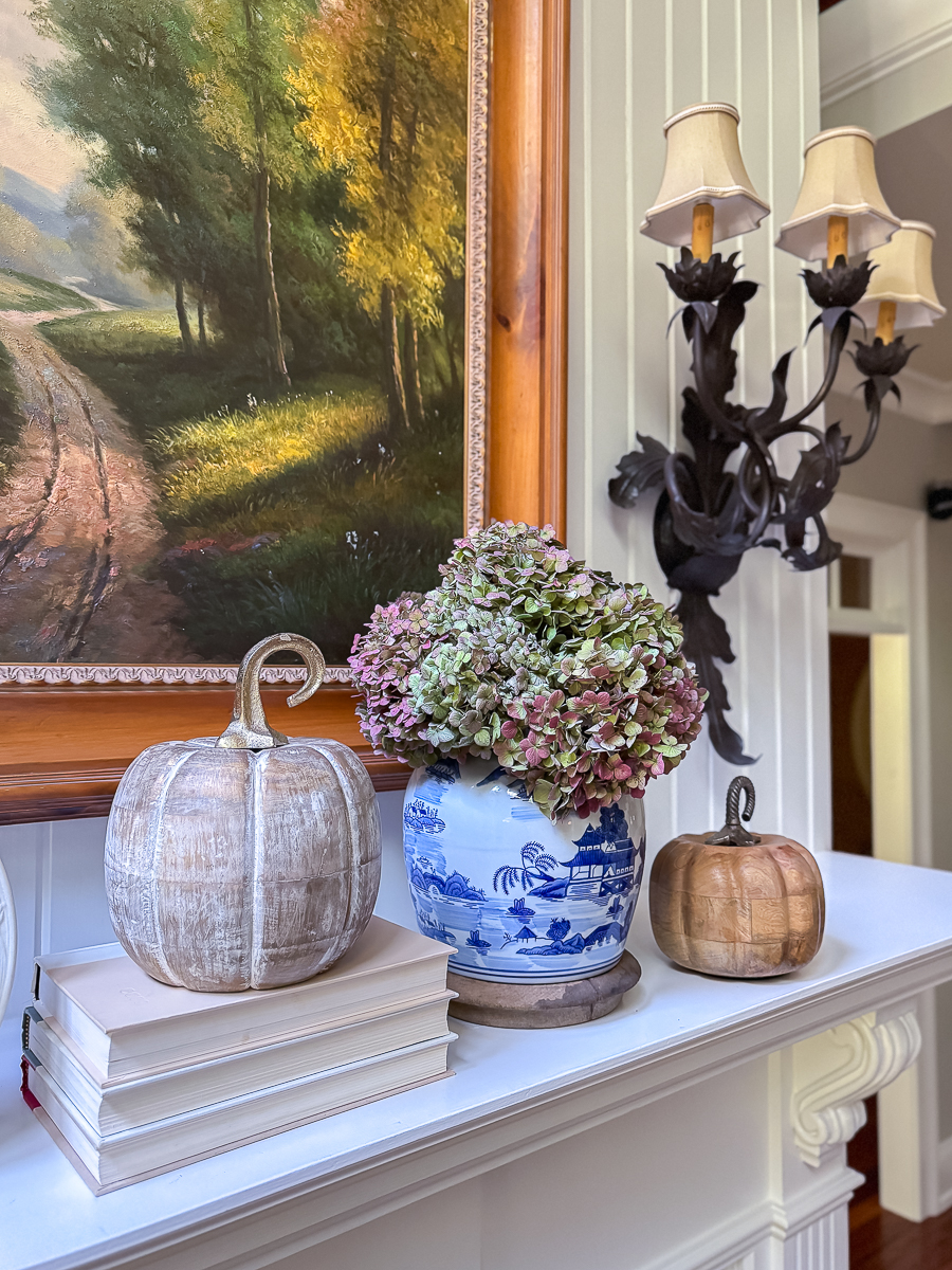 Stacked books, wooden pumpkins, and a chinoiserie vase with dried hydrangeas styled on a fall mantel.