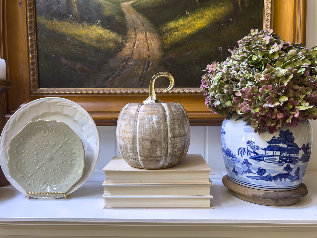 Decorative plate, stacked books, wooden pumpkins, and a chinoiserie vase with dried hydrangeas styled on a fall mantel.