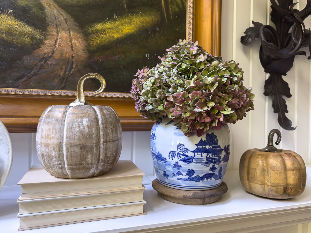 Stacked books, wooden pumpkins, and a chinoiserie vase with dried hydrangeas styled on a fall mantel.