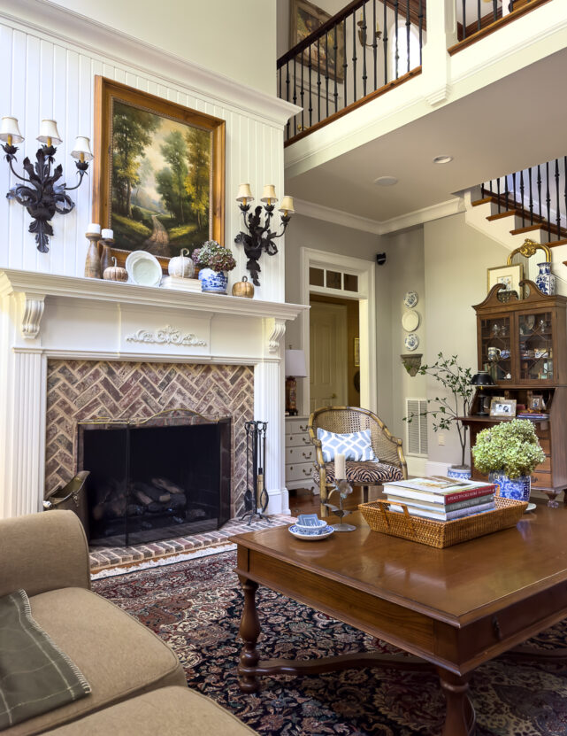 A cozy fall living room with a brick fireplace, classic artwork, and a decorated mantel featuring chinoiserie, pumpkins, and dried hydrangeas.