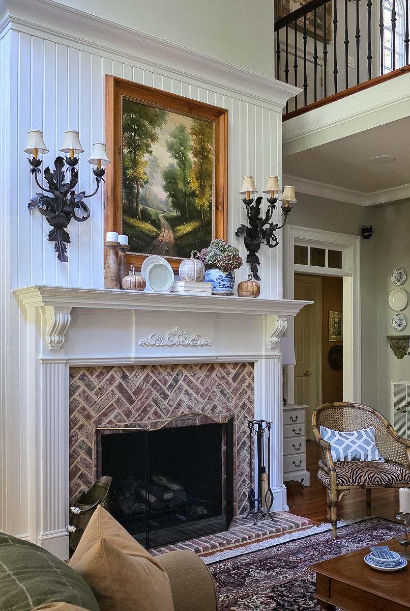 A cozy fall living room with a brick fireplace, classic artwork, and a decorated mantel featuring chinoiserie, pumpkins, and dried hydrangeas.