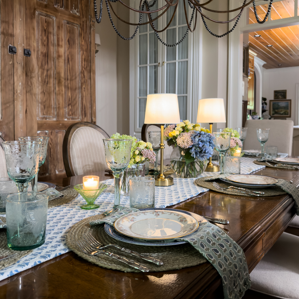 Collected summer tablescape with layered vintage dishes, woven placemats, blue glassware, and fresh hydrangeas.