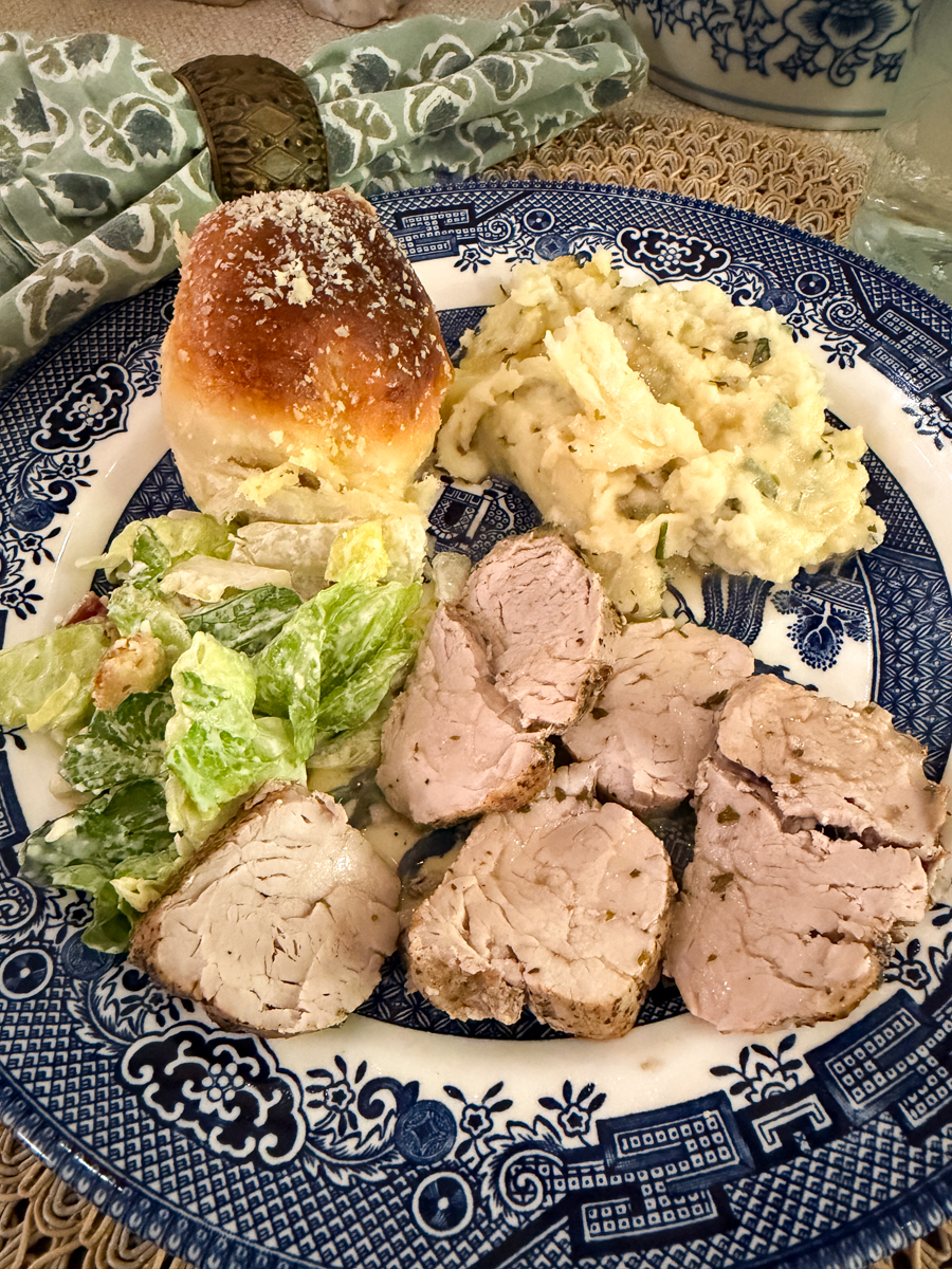 Sliced herb pork tenderloin served with mashed potatoes, Caesar salad, and a dinner roll on blue and white china