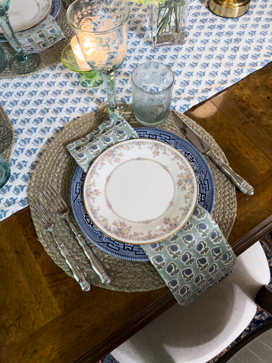 Layered summer place setting with vintage floral china, blue dinner plate, woven placemat, patterned napkin, and etched blue glassware.
