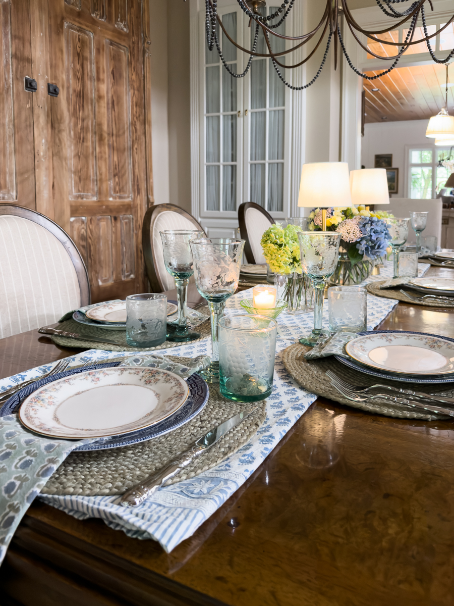 Simple layered summer tablescape with collected decor featuring vintage china, woven placemats, blue patterned table runner, floral arrangements, and cordless lamps.