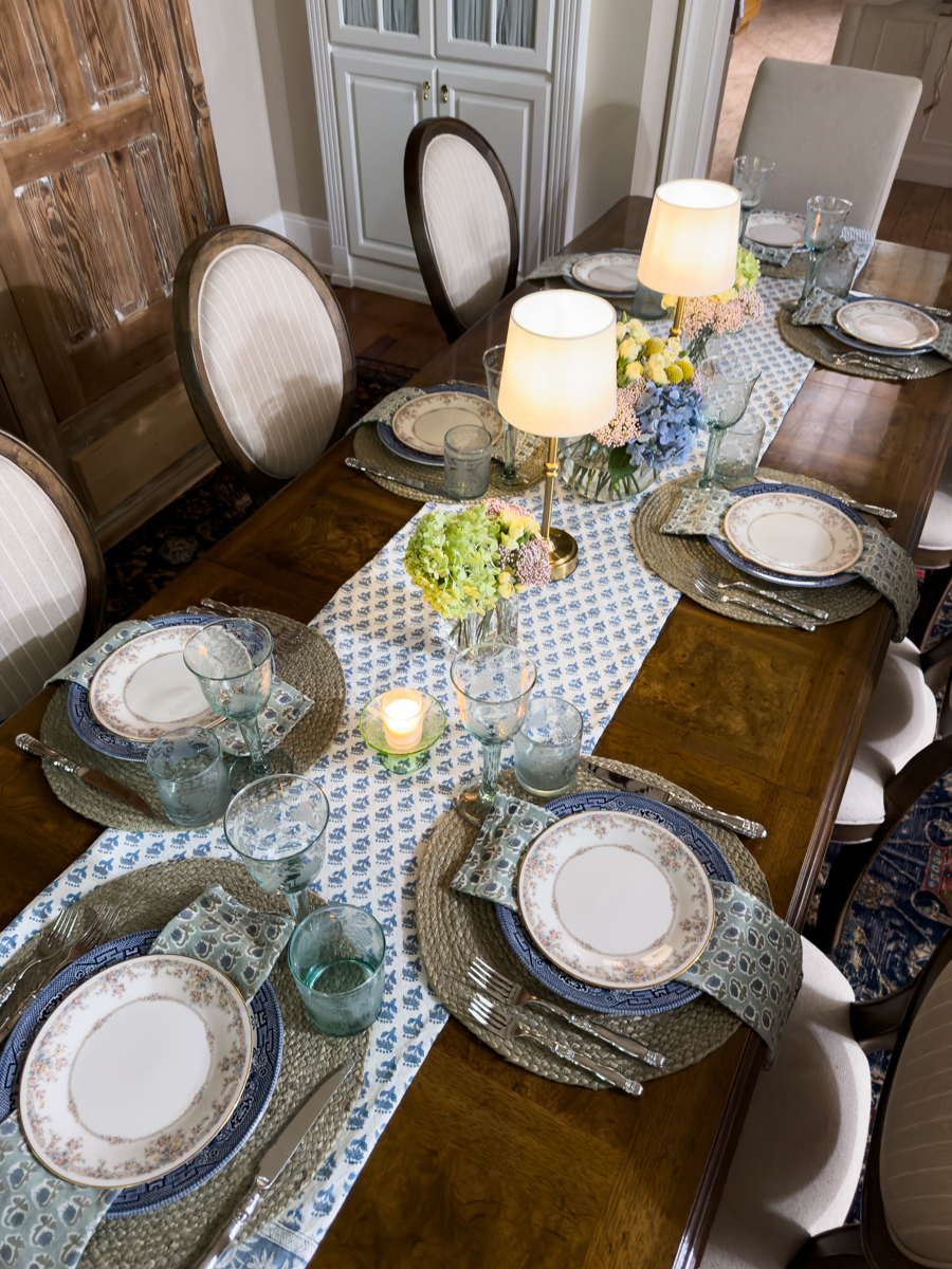 Overhead view of a layered summer dining table with collected decor, blue table runner, fresh flowers, and vintage tableware.