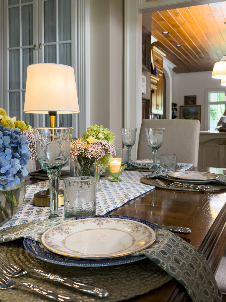 Side view of a layered summer tablescape with vintage china, blue linens, fresh flowers, and cordless table lamps.