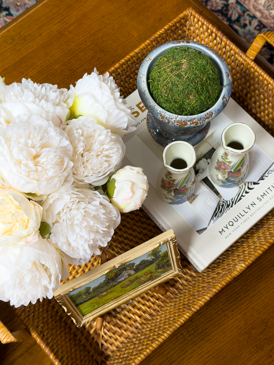 Wicker tray styled with flowers, moss ball, vintage sake pitchers, and books on a coffee table.