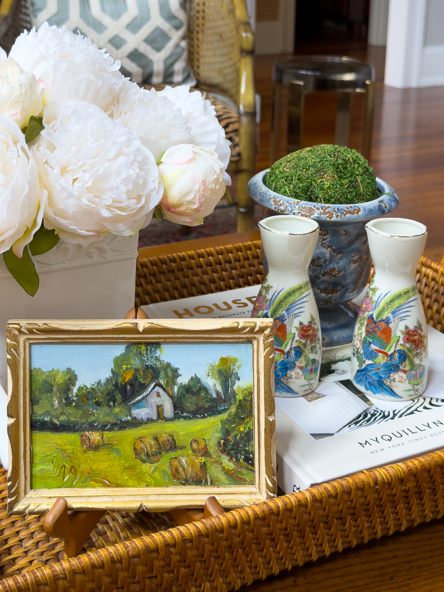 Small vintage landscape painting displayed inside a wicker tray on a coffee table.