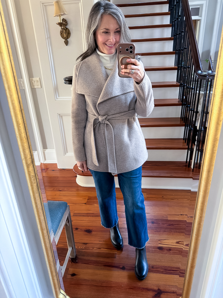 Woman wearing a neutral wrap coat with jeans and boots in a mirror selfie.