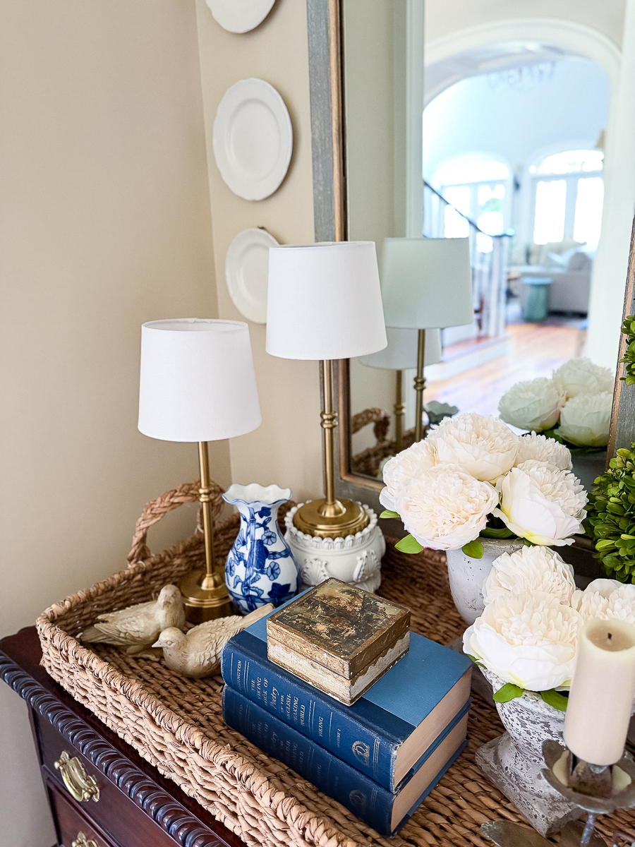 Vintage books stacked in woven tray with peonies and brass lamp for spring decor.